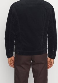 Veste en velours côtelé noir avec un empiècement au dos, des poignets à boutons et une coupe droite. Tissu texturé avec des détails de côtes verticales.