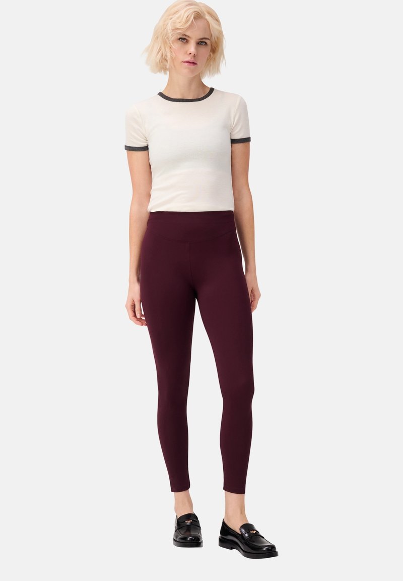 Witte T-shirt met korte mouwen en donkergrijze ribbels aan de hals, gecombineerd met bordeauxrode leggings en zwarte instappers. Zacht materiaal, strakke pasvorm.
