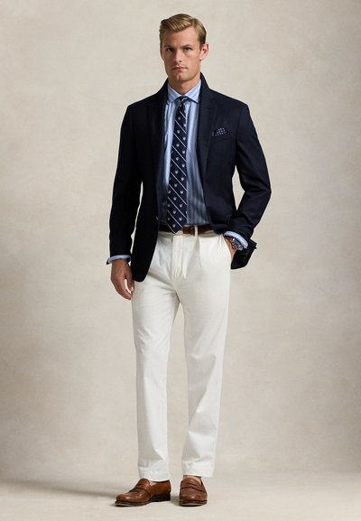 Mann, der eine marineblaue Jacke, ein blau gestreiftes Hemd, eine marineblaue gemusterte Krawatte, weiße Hosen, einen braunen Gürtel, braune Loafers und eine Armbanduhr trägt, vor einfachem Hintergrund steht.