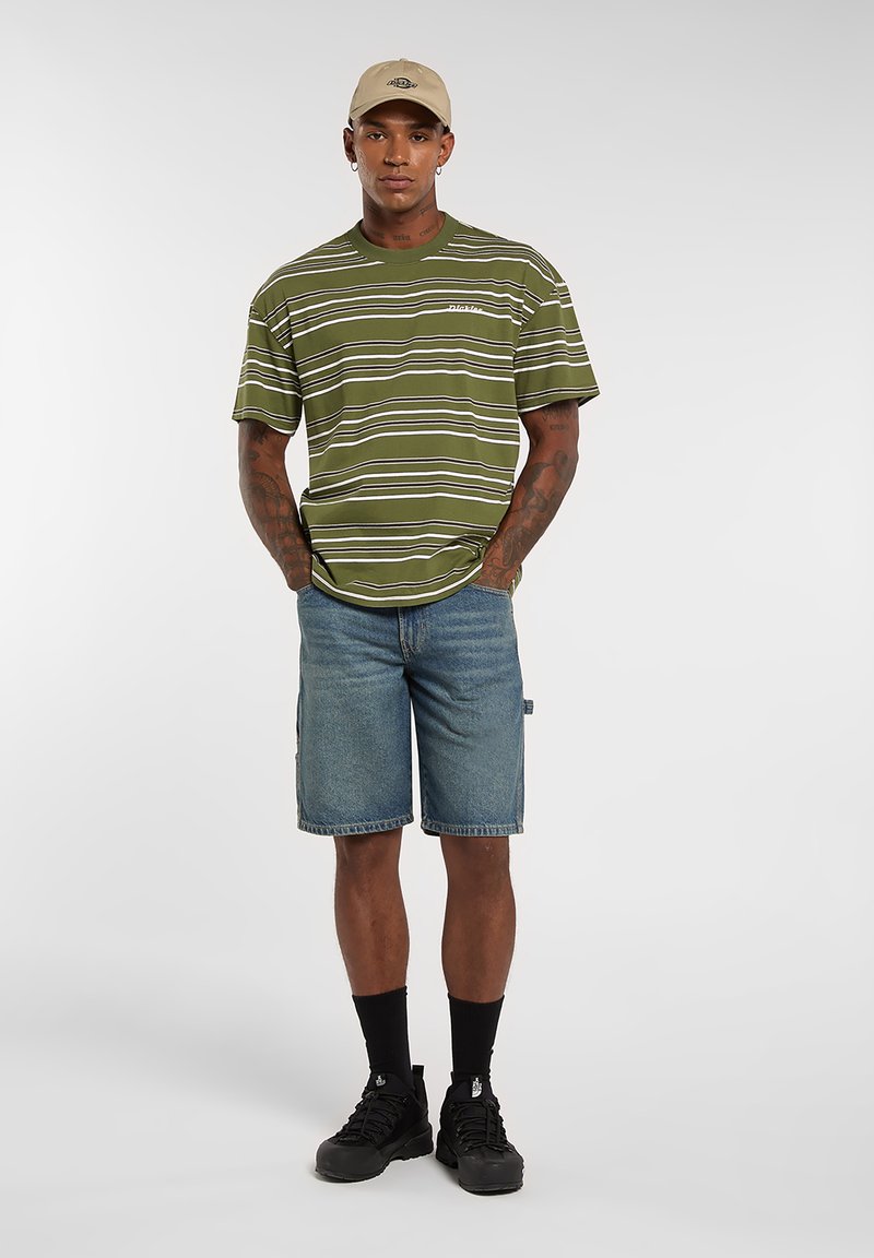 Uomo che indossa una maglietta a righe verde oliva, pantaloncini di jeans, cappello beige, calzini neri e scarpe da ginnastica nere in piedi con le mani nelle tasche.