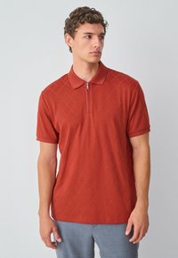Polo shirt couleur rouille avec col zippé, motif en diamant texturé, manches courtes et coupe décontractée. Associé à un pantalon gris clair.