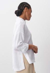 White linen shirt with a relaxed fit, featuring a collar, rolled-up sleeves, and side slits, displayed against a plain background.