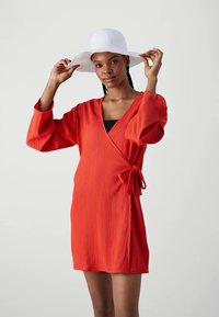 Vestido rojo envolvente con tela texturizada, mangas largas y un detalle de lazo en la cintura. Lleva puesto un sombrero blanco de ala ancha.