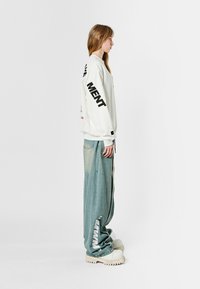 Sudadera blanca con texto negro en las mangas, combinada con unos pantalones de denim oversized en azul claro. Presenta piernas anchas y zapatillas blancas gruesas.