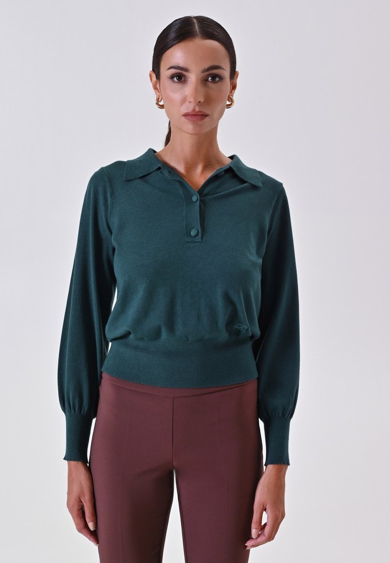 Pullover verde lavorato a maglia con colletto a polo, chiusura con bottoni e orlo a coste. Maniche lunghe con polsini aderenti. Indossato con pantaloni marroni a vita alta.