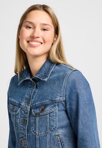 Jonge vrouw die glimlacht en een blauwe spijkerjasje draagt tegen een effen lichte achtergrond.