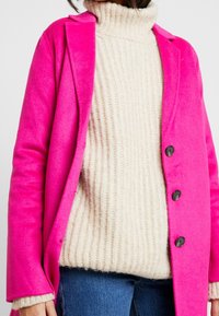 Blazer di lana fucsia con risvolti e tre bottoni neri, sovrapposto a un maglione a collo alto in maglia grossa crema e jeans blu.