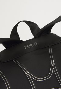 Fekete nylon táska ezüst varrási részletekkel, két szövetfogantyúval és egy címkével, amelyen a "REPLAY" felirat egy fekete folton van gravírozva.