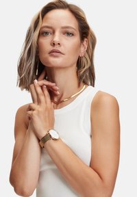 Goudkleurige horloge met een mesh band, witte wijzerplaat en minimalistisch ontwerp. Het model draagt een wit geribbeld topje en gouden hoepeloorbellen.