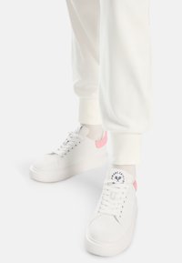 Weiße Sneakers mit einer strukturierten Oberseite, die rosa Akzente an der Ferse und ein rundes Logo auf der Zunge aufweisen. Kombiniert mit weißen Jogginghosen.