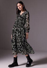 Lange zwart-witte jurk met patroon, V-hals, elastische taille en doorschijnende mouwen, gecombineerd met kniehoge zwarte laarzen.
