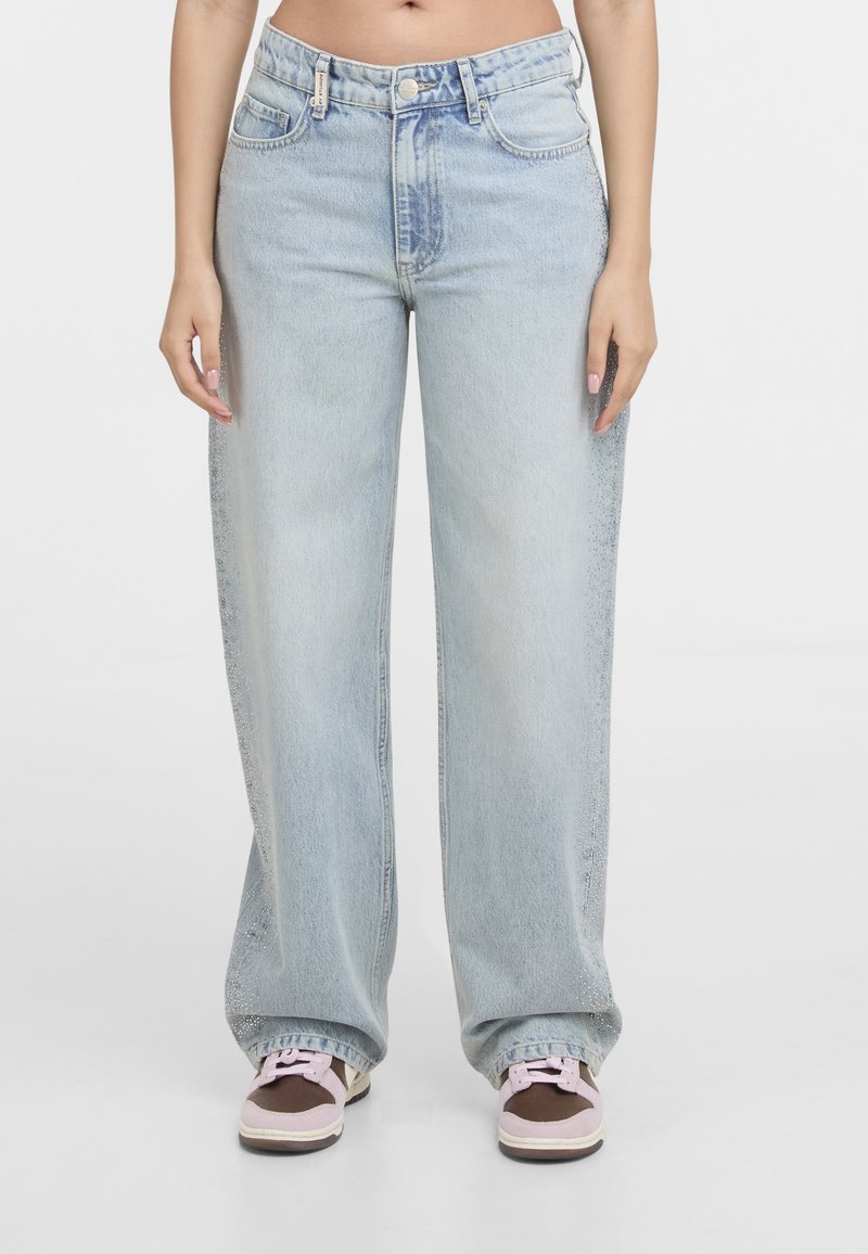 Personne portant un jean bleu clair ample et des baskets marron et roses, debout devant un fond blanc.