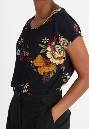 Femme portant un chemisier à manches courtes fleuri bleu marine foncé, rentré dans un pantalon noir taille haute, une main dans la poche.