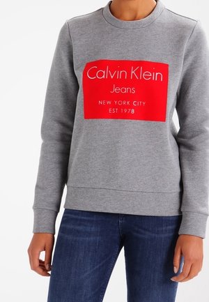 Osoba nosząca szarą bluzę z czerwonym prostokątnym logo z napisem „Calvin Klein Jeans New York City Est 1978” oraz ciemnoniebieskie dżinsy.