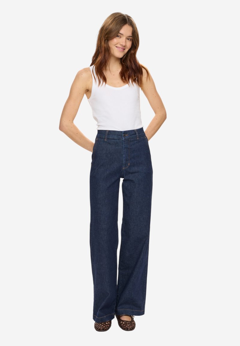 Femme aux longs cheveux bruns portant un débardeur blanc, un jean bleu taille haute à jambes larges, et des chaussures tressées marron, souriante avec les mains derrière le dos.