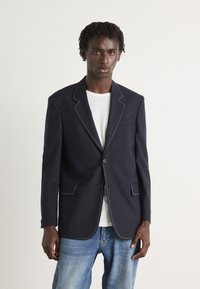 Blazer azul-marinho com costura branca em contraste, fecho de um único botão, lapelas recortadas e dois bolsos frontais, usado sobre uma t-shirt branca.