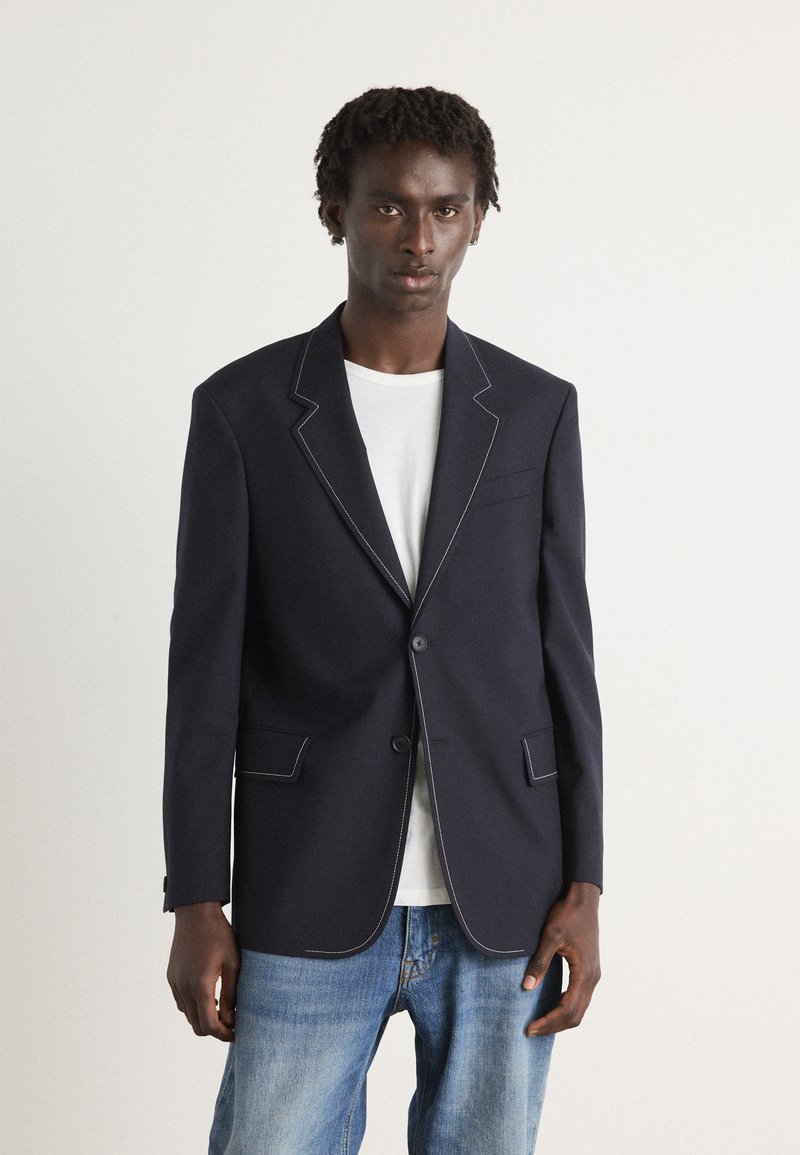 Blazer azul-marinho com costura branca em contraste, fecho de um único botão, lapelas recortadas e dois bolsos frontais, usado sobre uma t-shirt branca.