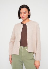 Ελαφρύ μπεζ ζακέτα με στρογγυλή λαιμόκοψη, διακριτική υφή, κουμπιά μπροστά και χαλαρή γραμμή στους ώμους, συνδυασμένη με καφέ τοπ και πράσινο παντελόνι.