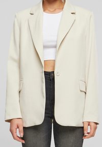 Světle béžový blazer vyrobený z měkké tkaniny, se zářezovým límcem, zapínáním na knoflíky a dvěma předními kapsami. Nosič má na sobě bílý crop top.
