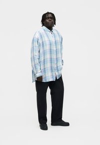 Hombre con rastas que lleva una camisa de cuadros azul claro de gran tamaño, pantalones negros y zapatos negros, de pie contra un fondo blanco.