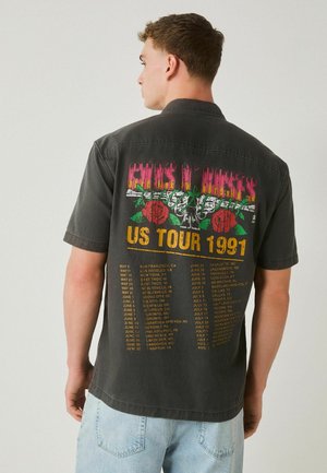 Mann trägt schwarzes Guns N' Roses US Tour 1991 Shirt mit roten Rosen, Waffen-Grafik und gelben Tourdaten auf der Rückseite.