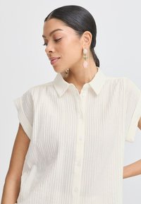 Camicia plissettata di colore chiaro con colletto e maniche corte. Il tessuto è testurizzato e presenta bottoni lungo la parte anteriore.