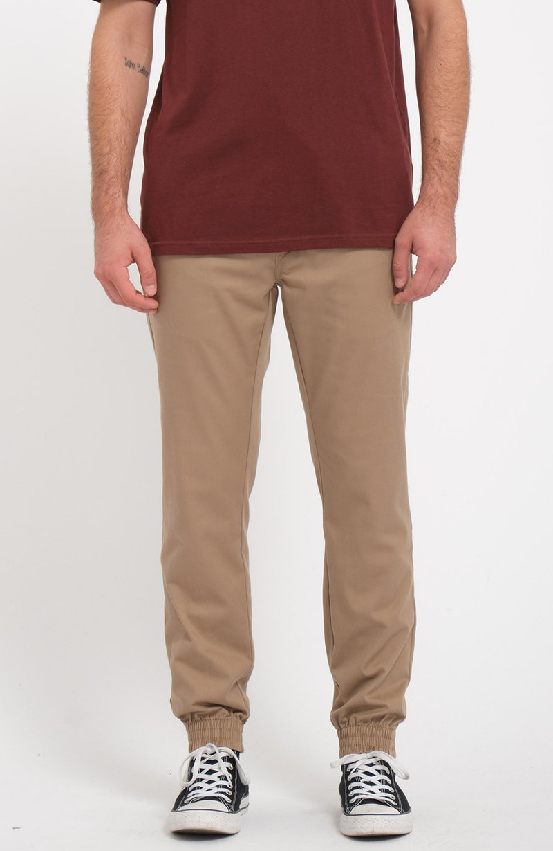 Pantalons beiges fuselés avec des poignets élastiques, en coton, poches latérales, portés avec des baskets noir et blanc, associés à une chemise bordeaux.