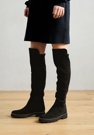 Μαύρες σουέτ μπότες πάνω από το γόνατο με επίπεδο, ραβδωτό πάτο και χρυσές καρφίτσες κατά μήκος της άκρης. Λείος υφάς και εφαρμοστό σχέδιο.