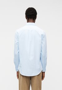 Άνδρας με τη πλάτη, φορώντας ανοιχτό μπλε μακρυμάνικο πουκάμισο και μπεζ παντελόνι μπροστά σε απλό λευκό φόντο.