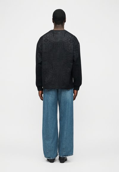 Άνδρας με την πλάτη, φορώντας μαύρο μακρυμάνικο πουκάμισο με λεπτό μοτίβο και μπλε τζιν παντελόνι με φαρδιά μπατζάκια, μπροστά σε απλό φωτεινό φόντο.