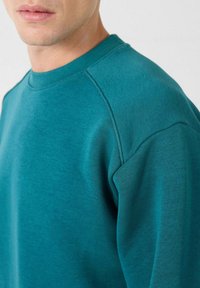 Teal sweatshirt met een ronde hals en raglanmouwen. Gemaakt van zachte, textuurstof met een gladde afwerking. Close-up van de schouder.
