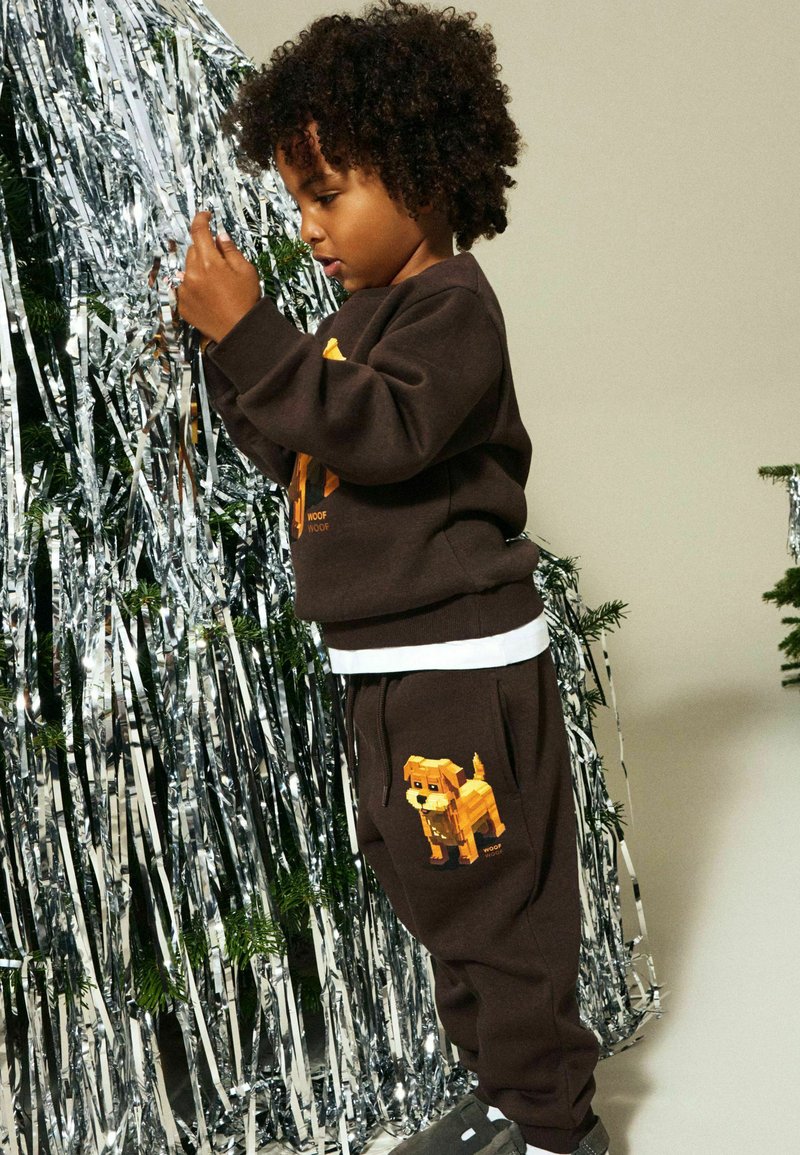 Brun genser og joggebukser med et pikselert design av en gul hund. Stoffet ser mykt ut; antrekket har en hvit midjebåndsdetalj.