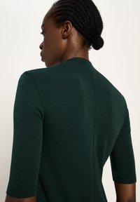Woman with braided hair bun wearing a dark green fitted top with three-quarter sleeves, shown from the back against a plain background.