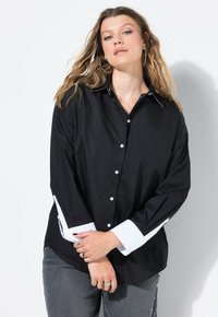 Giovane donna con capelli lunghi e mossi che indossa una camicia nera con bottoni e dettagli bianchi sui polsini e sul colletto, abbinata a jeans grigi e orecchini a cerchio argentati.