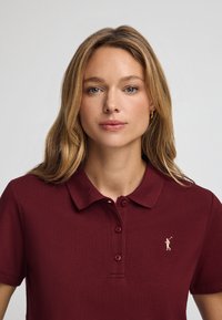 Burgundy pikétröja med krage, tre knappar och en broderad logotyp. Tillverkad av texturerad tyg, med korta ärmar.