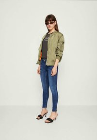 Olivo zelena bomber jakna z zadrgo, kombinirana s črno majico z grafiko in modrimi oprijetimi kavbojkami. Črne sandale s peto poudarjajo videz.