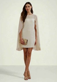 Γυναίκα που φοράει ένα μπεζ φόρεμα-κάπα, κρατώντας μια υφασματική χρυσή τσάντα, σε συνδυασμό με χρυσά ψηλοτάκουνα σανδάλια, με φόντο σε ουδέτερο χρώμα.