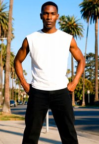 Moški stoji na prostem z rokami v žepih, oblečen v belo brezrokavnik in črne hlače, v ozadju palmova drevesa in modro nebo.