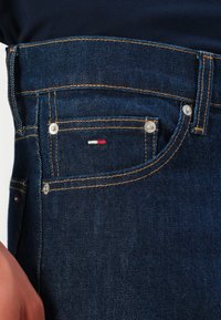 Poche d'un jean en denim bleu foncé avec des coutures jaunes, des rivets en métal et un petit logo brodé rouge, blanc et bleu près du bord supérieur.