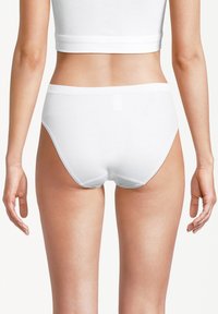 Femme portant des culottes blanches en coton uni et un débardeur blanc court, vue de dos sur un fond clair.