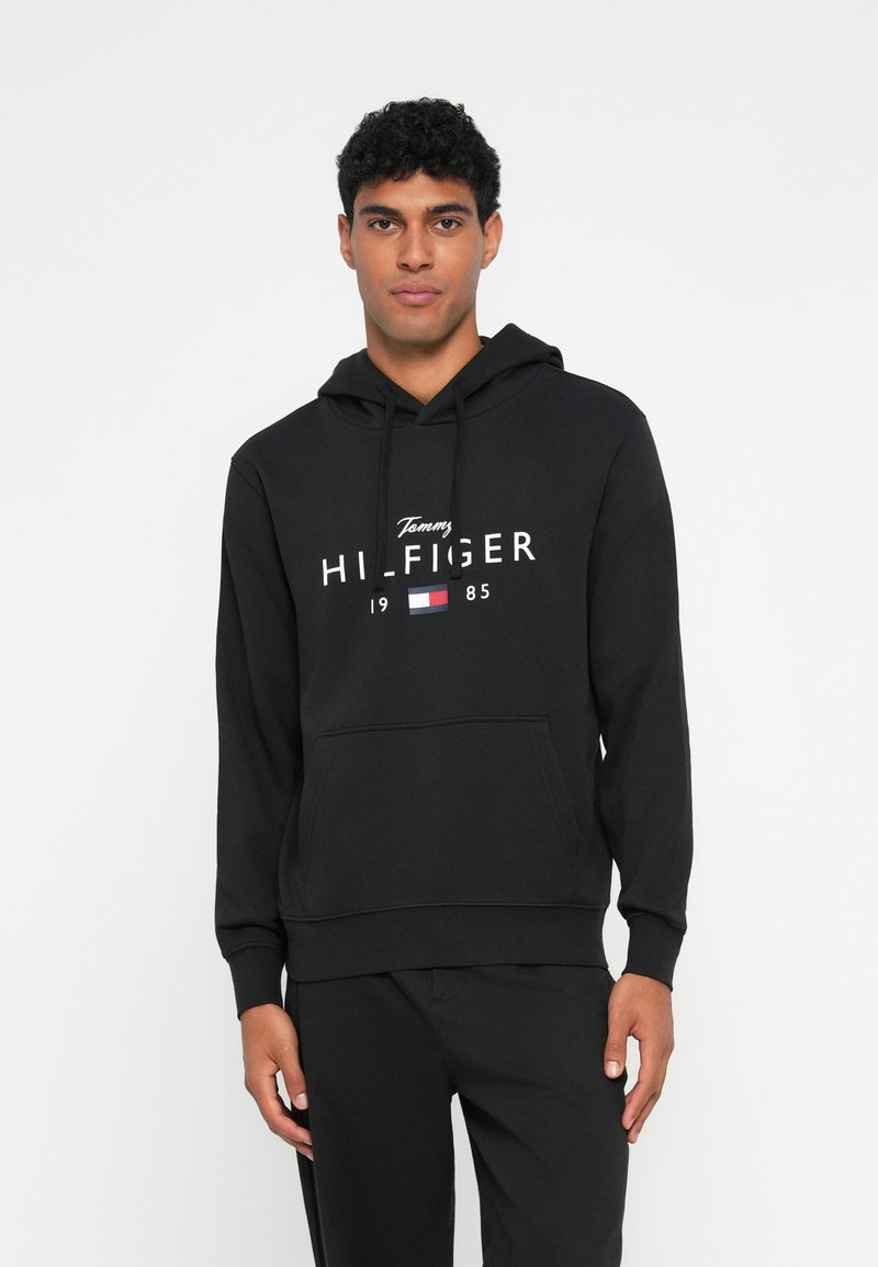 Sweatshirt preto feito de mistura de algodão, com bolso frontal tipo canguru, capuz com cordão e texto de marcação em branco com acentos em vermelho e azul.