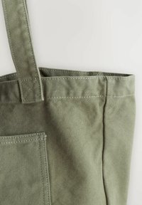 Olive green cotton tote bag with a textured surface, featuring a single short handle and a front pocket, reinforced stitching.
