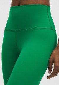 Zaļas augstās sporta legins, kas izgatavoti no elastīga, gluda materiāla; tām ir plakanas šuves un pieguļošs dizains, kas nodrošina komfortu un elastību.