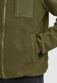 Olivroheline fliisjacket, millel on lukuga esikülg, tekstureeritud viimistlus, suur rakendustasku ja küljetaskud.