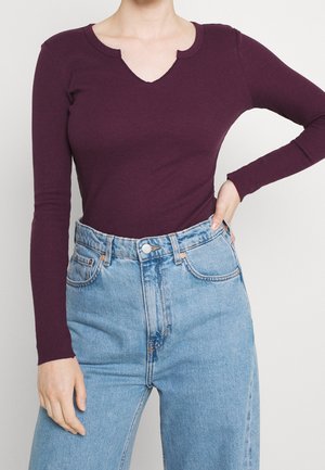 Personne portant un haut à manches longues, côtelé, violet foncé avec une encolure fendue et un jean bleu clair taille haute, main sur la hanche.
