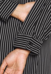 Bras gauche plié derrière le dos, portant une chemise noire à fines rayures verticales blanches et poignet boutonné.