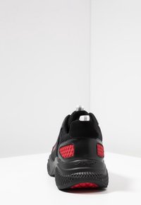 Schwarzer Sportschuh mit roten Mesh-Details, strukturiertem Sohle und weißen Zuglaschen. Verfügt über ein schlankes Design und ein auffälliges Markenlogo an der Ferse.