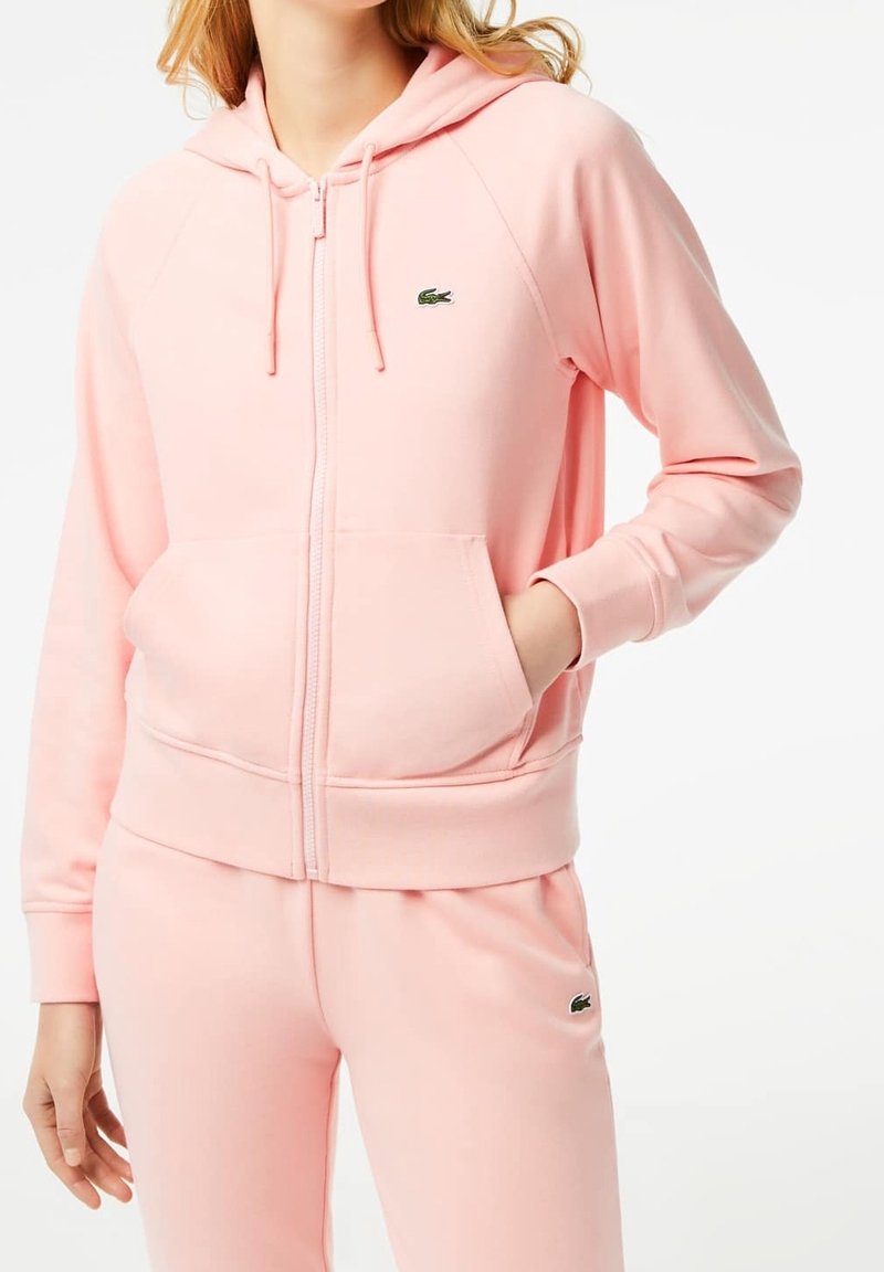 Rosa zip-up hoodie tillverkad av mjukt tyg. Har en huva, framfickor och en liten grön logotyp på bröstet. Slät textur, avslappnad passform.