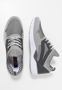 Zapatillas deportivas grises con una parte superior de malla estampada, que presenta rayas y una superposición de plástico, y una suela de goma blanca.