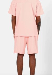 Camicetta rosa chiaro a maniche corte e pantaloni corti abbinati in un tessuto strutturato, con una vestibilità ampia e design semplice. Nessun logo o motivo visibile.